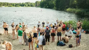 Cilvēku grupa stāv smilšainā pludmalē un skatās uz ezeru, kurā redzami airu dēļu braucēji.