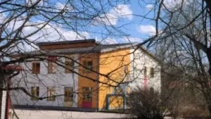 Divstāvu skolas ēka ar oranžām un baltām sienām, ko daļēji aizsedz koku zari pavasarī.