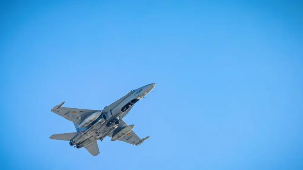 Pelēka militārā iznīcinātājlidmašīna lido skaidrās, gaiši zilās debesīs no apakšas skatoties.