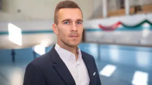 Vīrietis tumšā žaketē un baltā kreklā pozē peldbaseina fonā ar izplūdušu skatu.