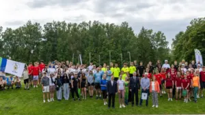 Liela jauniešu grupa ar amatpersonām stāv uz zaļas zāles pretī mežam kopīgā bildē.
