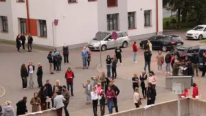 Cilvēku grupa pulcējas bruģētā laukumā pie daudzdzīvokļu ēkas, pētot kartes un sazinoties savā starpā.
