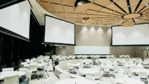 Plaša konferenču zāle ar apaļiem galdiem un lieliem ekrāniem pasākuma norisei.
